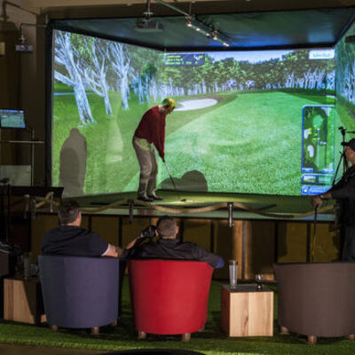 Bild vergrößern: Blick auf den Indoorgolf-Simulator mit einem Spieler und Zuschauern